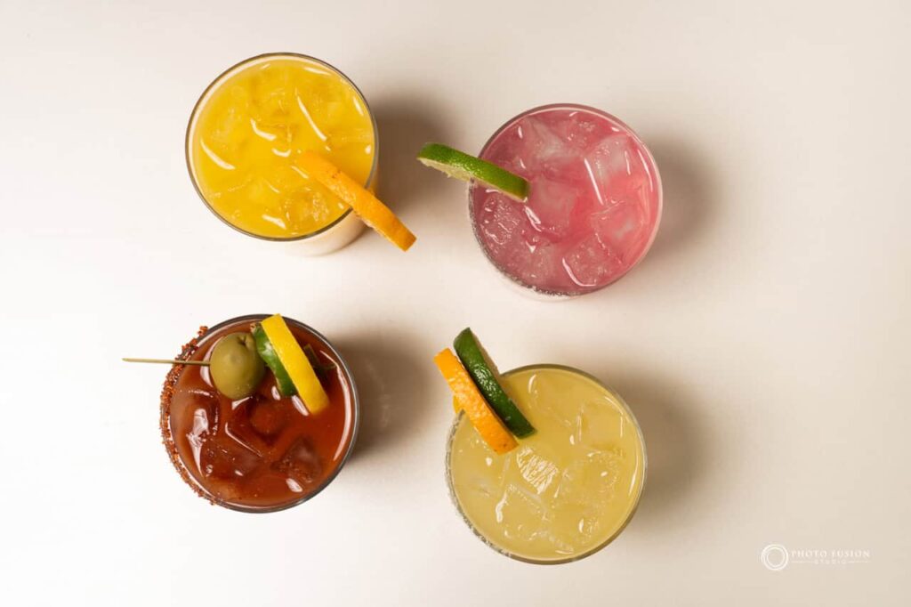 This image is a top down shot of four drinks. The top left is an orange drink with an orange slice, the top right is a pink drink with a lime slice, the bottom right is a orange drink with both a lime and orange slice, and the bottom left is a red drink with an olive, and lemon slice. This image was taken for the Jojo's Coffeehous eBreakfast & Brunch main menu