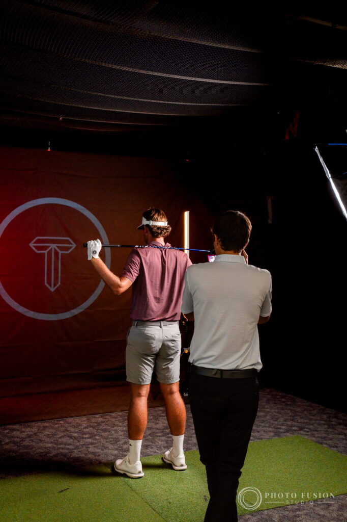An ASU golfer rests a club on his shoulder beside a True Spec fitter in front of a red True Spec logo backdrop