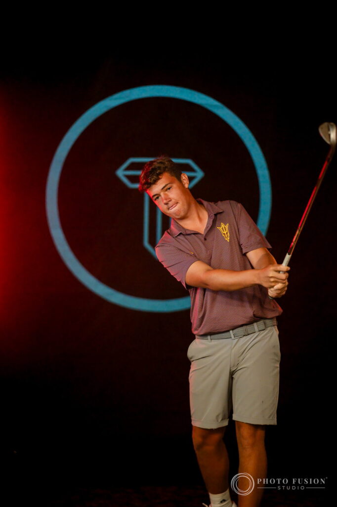 An ASU men's golf player finishes his swing in front of a glowing teal True Spec Golf logo backdrop