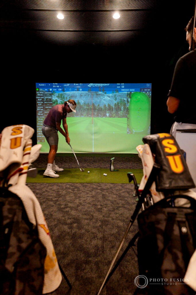 An ASU golfer hits an iron toward a Foresight simulator screen, framed by ASU headcovers on a bag