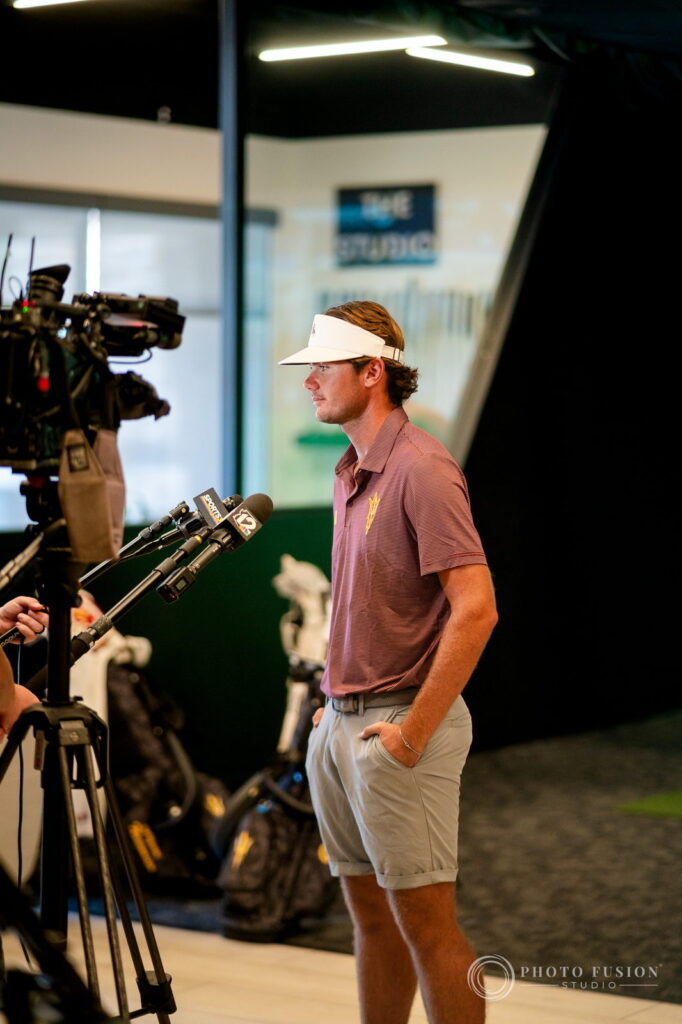 An ASU golfer in a white visor stands in front of TV microphones and a broadcast camera at True Spec Golf
