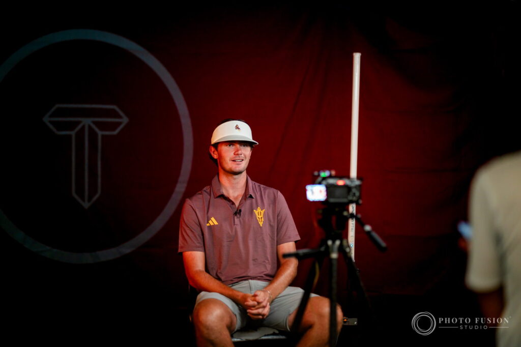 An ASU men's golf player sits for a filmed interview in front of a dark red True Spec Golf backdrop