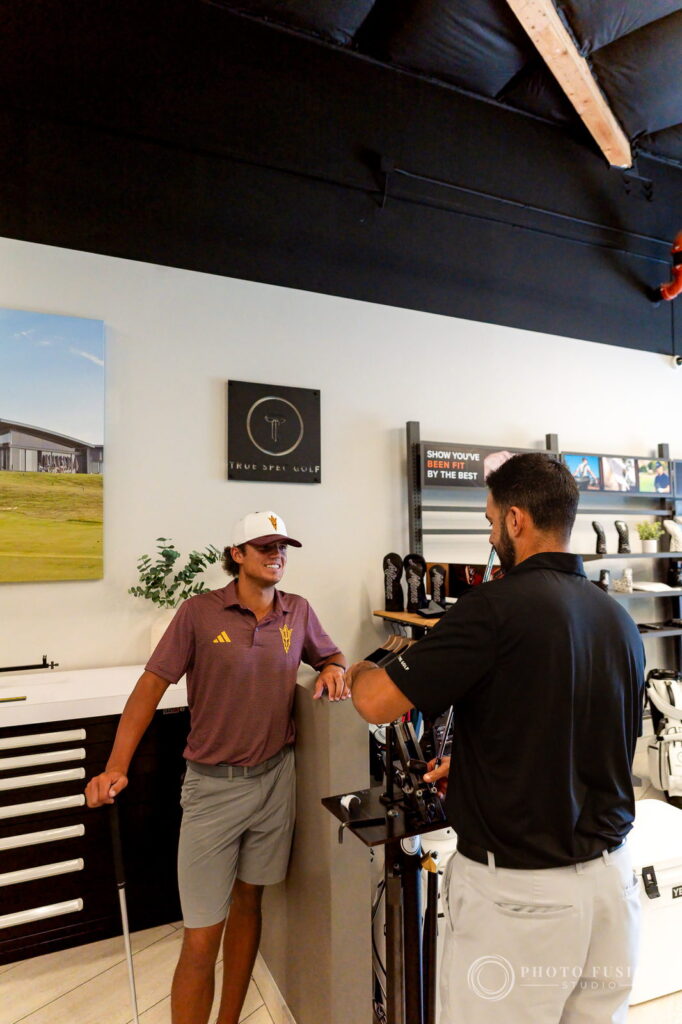 An ASU golfer leans on the counter while a True Spec fitter sorts shafts under a Been Fit By The Best sign