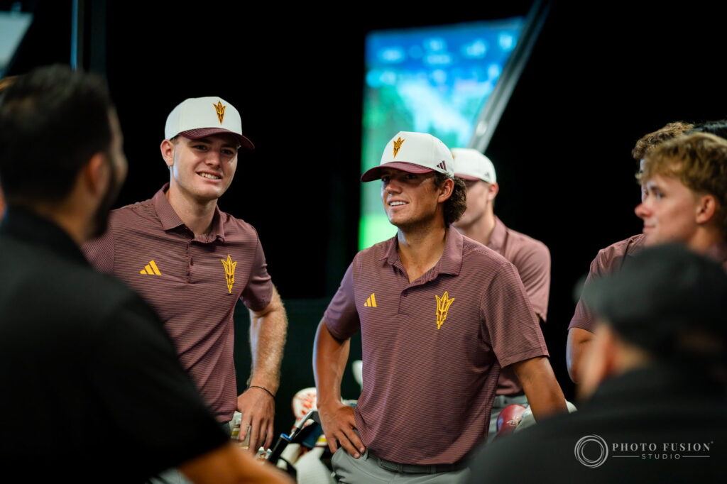 Two ASU men's golf players in maroon polos smile inside a fitting bay with simulator screens behind them