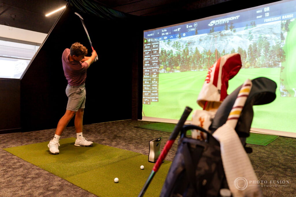 An ASU golfer swings in a True Spec Golf simulator bay with a golf bag and headcovers in the foreground