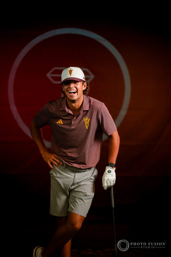 An ASU men's golf player laughs while leaning on his club in front of a dark red True Spec Golf backdrop