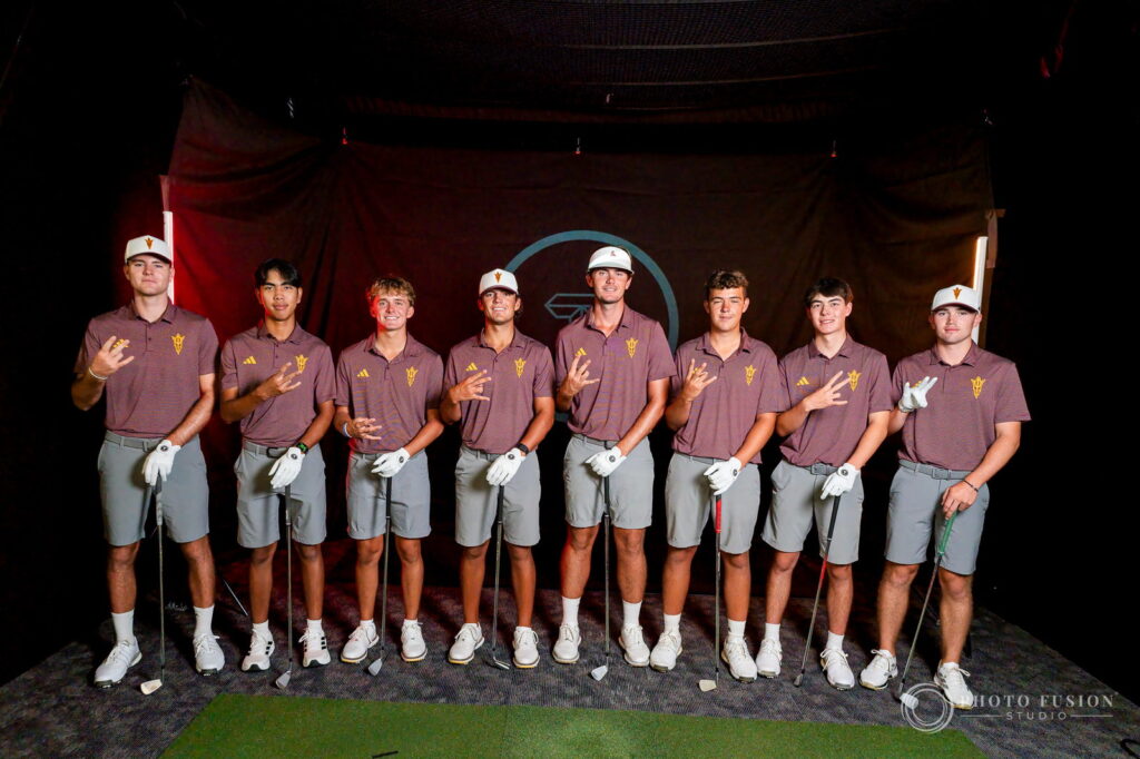 Eight ASU men's golf players in maroon polos raise the pitchfork sign inside a True Spec Golf hitting bay