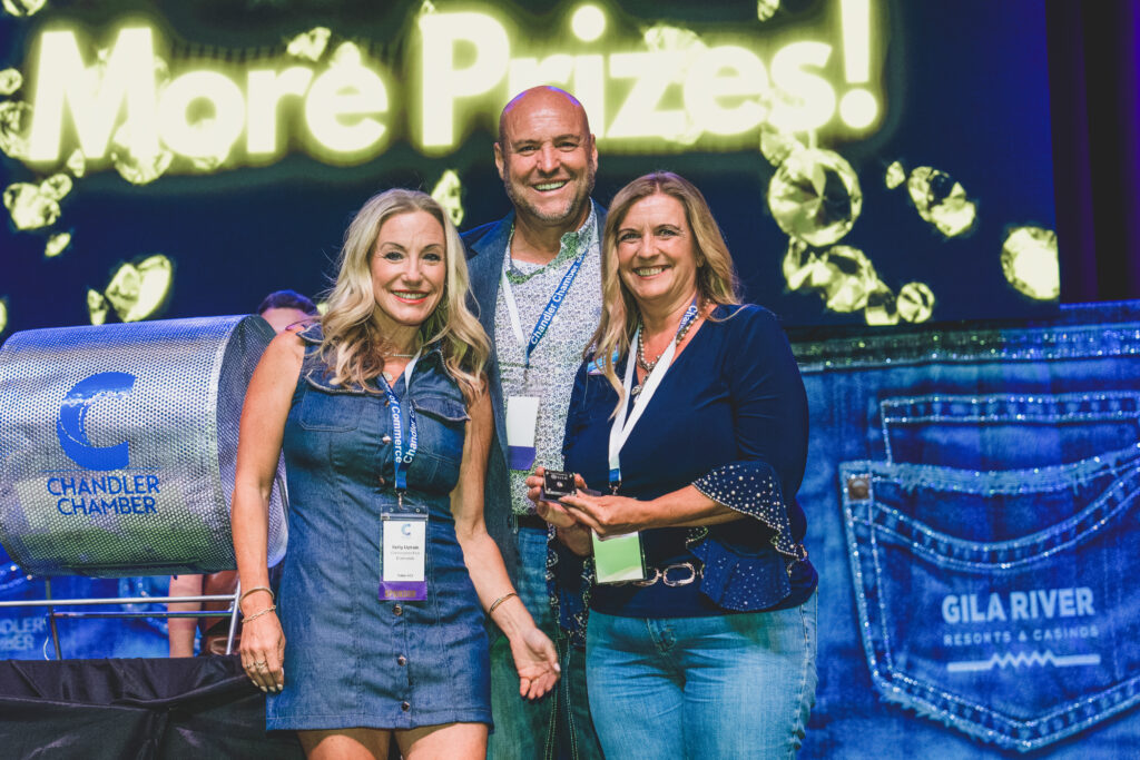 Three guests pose by a raffle drum in front of a More Prizes neon diamond screen at the Chandler Chamber Awards