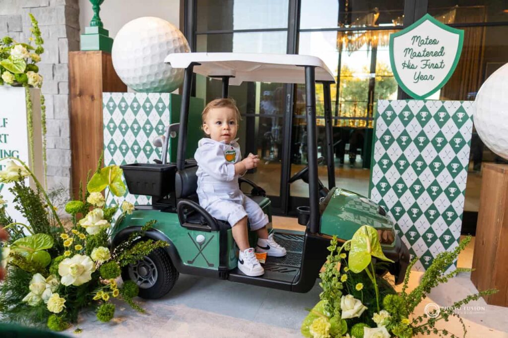 A child is sitting in his mini golf cart that is made for kids and smiling. He is celebrating his first birthday that is a masters golf theme party. There are flowers on both sides of the cart and giant golf ball displays as well.