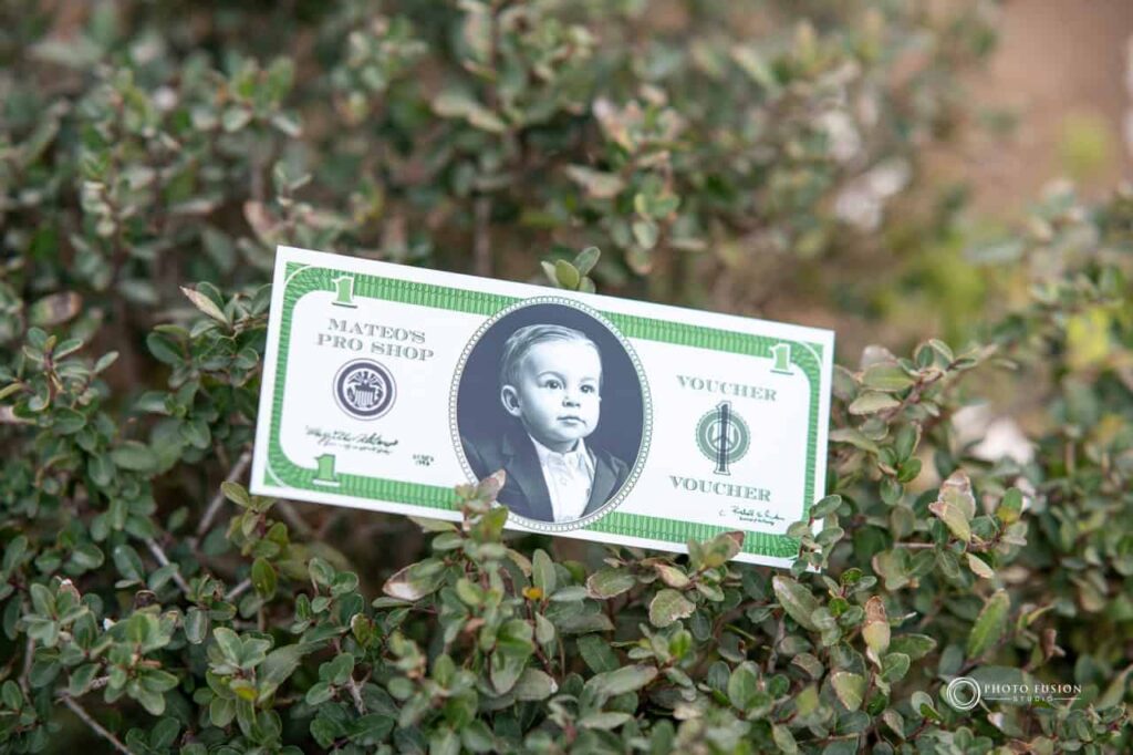 This is a photo of custom play money with a childs face on it. He is turing one and the money is a one dollar bill. It is in a green bush.