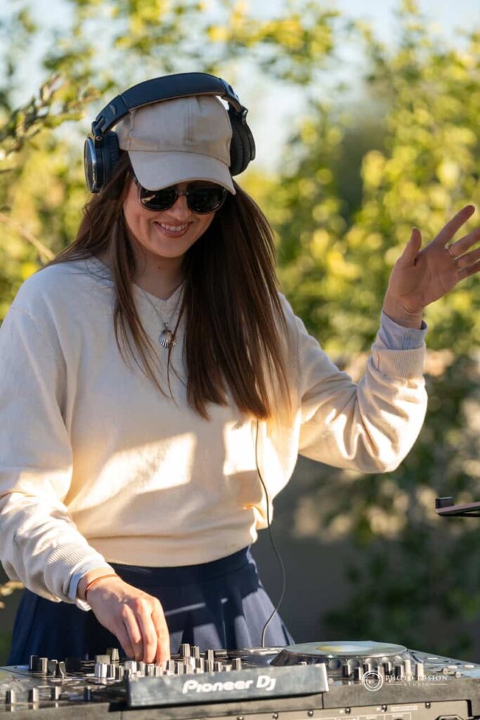 DJ at a one year olds birthday party. She has her headphones on and one hand in the air with theother turning the dials on the DJ board. She has a smile on her face as she works.