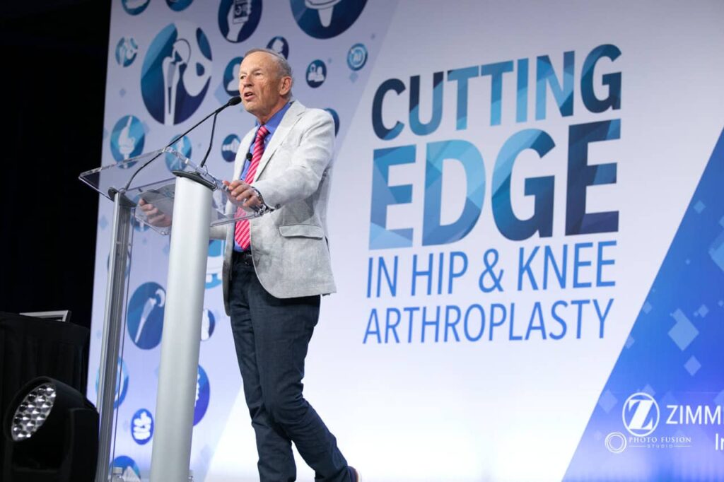 Man Standing on a stage while talking and giving a presentation to a group of people in a confrence room. The sign behind him says Cutting Edge in hip & knee arthroplasty