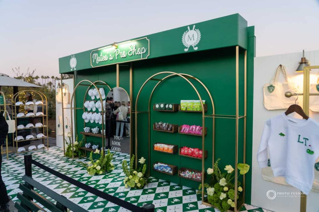 Photo of a mini pro shop at a one year old's birthday party that is masters golf theme. There is a mirror in the middle that says you look good on it. On the left side of the mirror there are hats hanging on the wall and golf balls on shelves under them. On the right there are snacks and candy on shelves hanging on the wall. On the far left of the wall there are tote bags hanging on the wall along with a shelf that thas vizors on it. The top of the pro shop has a sign that says Mateo's Pro Shop.