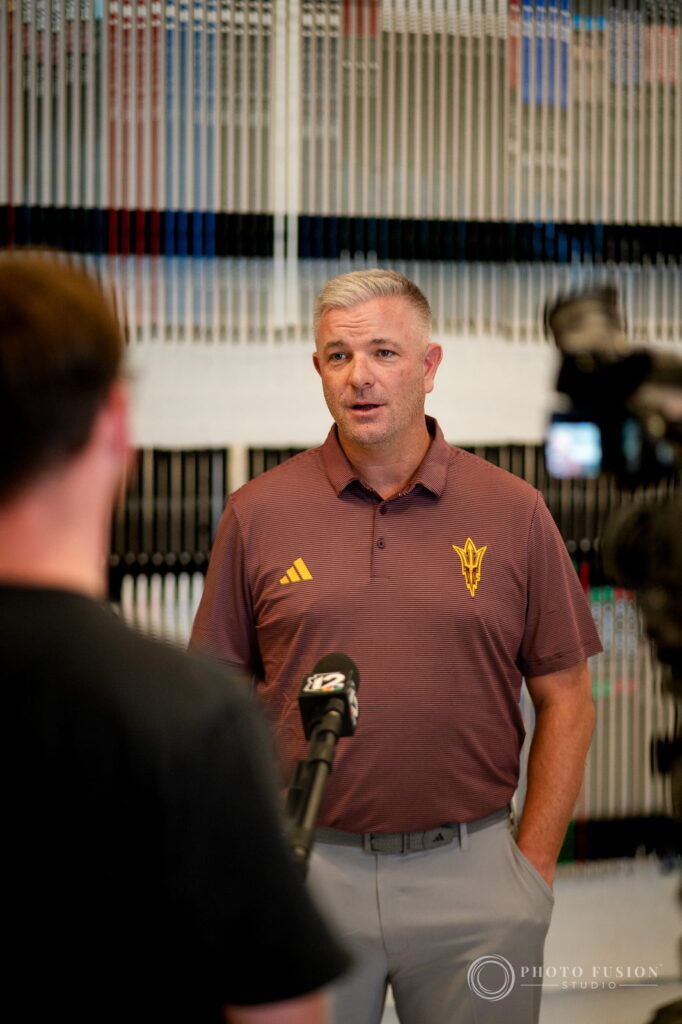 ASU men's golf head coach Matt Thurmond speaks to a TV reporter in front of a wall of golf shafts