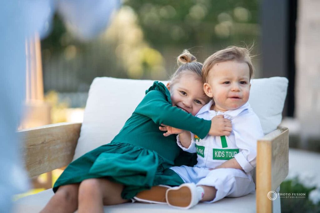 There are two kids in this image an the girl is hugging the boy, who is her younger borhter. It is his birthday and they are getting a sibling photo together. She is looking at the camera with a big smile on her face while hugging her younger brother. He is looking off into the distance.