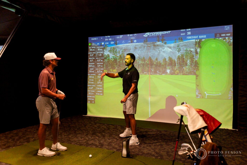A True Spec Golf fitter points at launch monitor data on the simulator screen while an ASU golfer listens