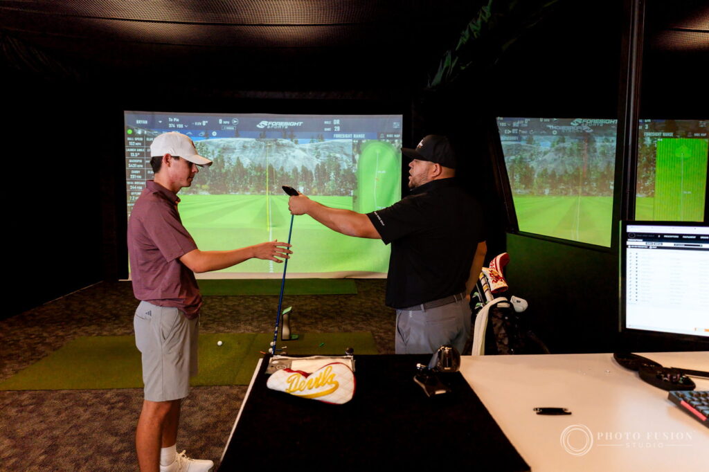 A True Spec Golf fitter hands a club to an ASU golfer in front of simulator screens showing swing data