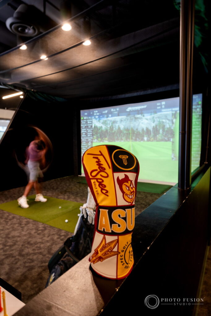 An ASU True Spec Golf headcover in the foreground with a blurred golfer swinging toward a simulator screen