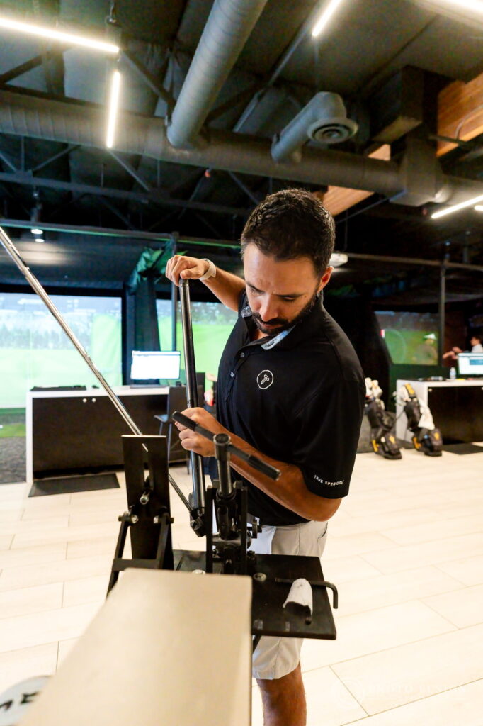 A True Spec Golf builder works a club at the shop's build press with the fitting bays behind him