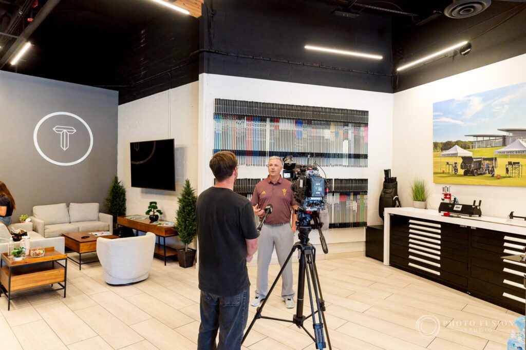 Wide view of the True Spec Golf Scottsdale showroom with a videographer filming a coach interview by the shaft wall