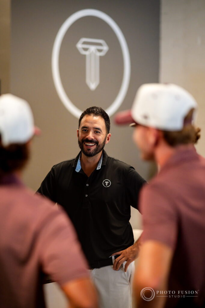 A True Spec Golf fitter in a black polo smiles while talking with ASU players in front of the True Spec logo