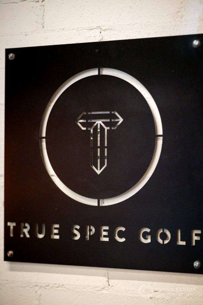 A brushed metal True Spec Golf sign mounted on a white painted brick wall