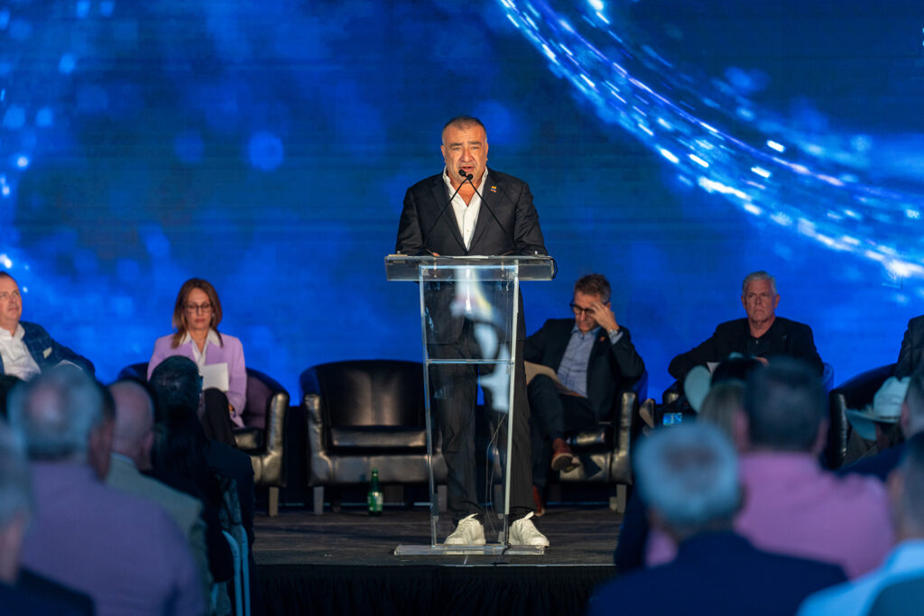 XNRGY CEO Wais Jalali at the podium addressing a seated audience with company dignitaries on stage behind him at the Mesa Gateway ribbon cutting.