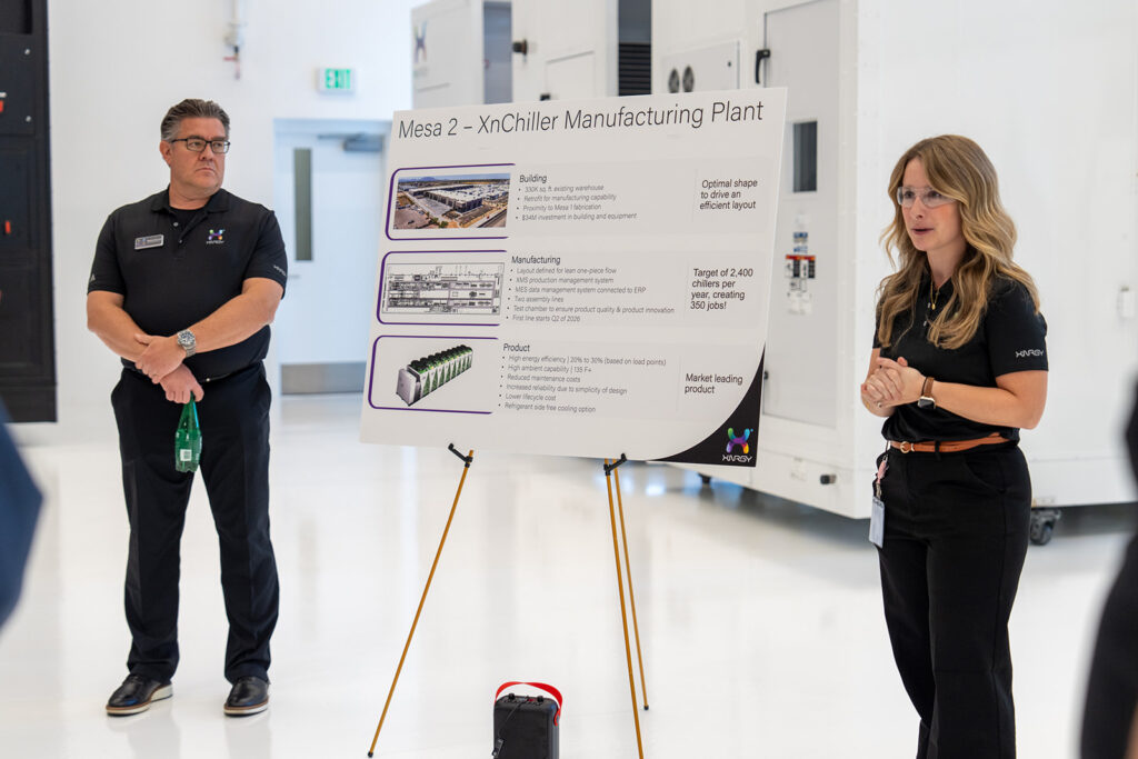Two XNRGY team members presenting the Mesa 2 XnChiller Manufacturing Plant information board with plant details and targets at the ribbon cutting open house.