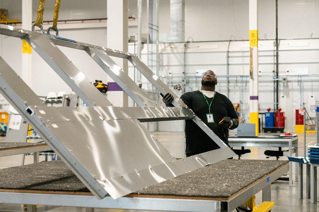 XNRGY worker in safety glasses lifting and positioning a large aluminum frame panel on an assembly worktable at the Mesa Gateway facility.