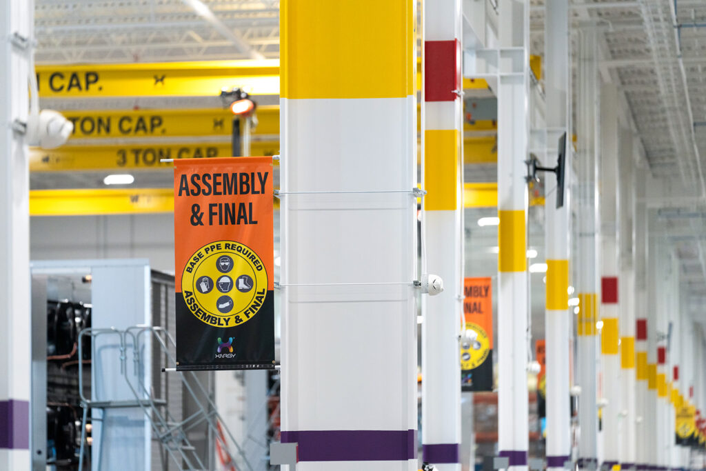 Bright orange ASSEMBLY & FINAL safety banners hanging from steel columns marking the PPE-required zone at the XNRGY Mesa Gateway facility.