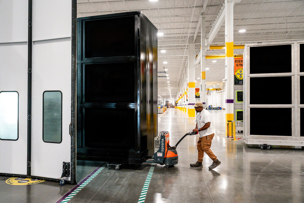 XNRGY worker moving a large black equipment cabinet through the production aisle with an electric pallet jack at the Mesa Gateway facility.