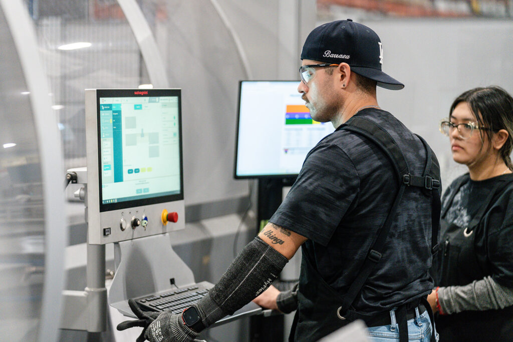XNRGY operator at a CNC sheet metal machine touchscreen control terminal with another team member observing at the Mesa Gateway facility.