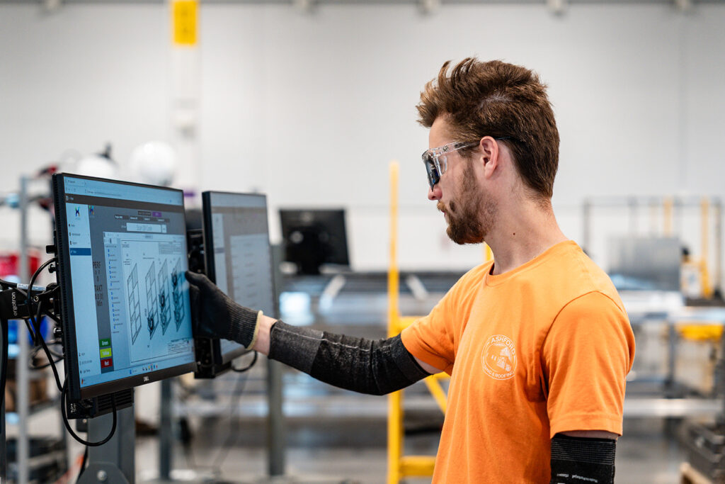 XNRGY worker in safety glasses reviewing engineering CAD drawings on dual monitors at a production workstation inside the Mesa Gateway facility.