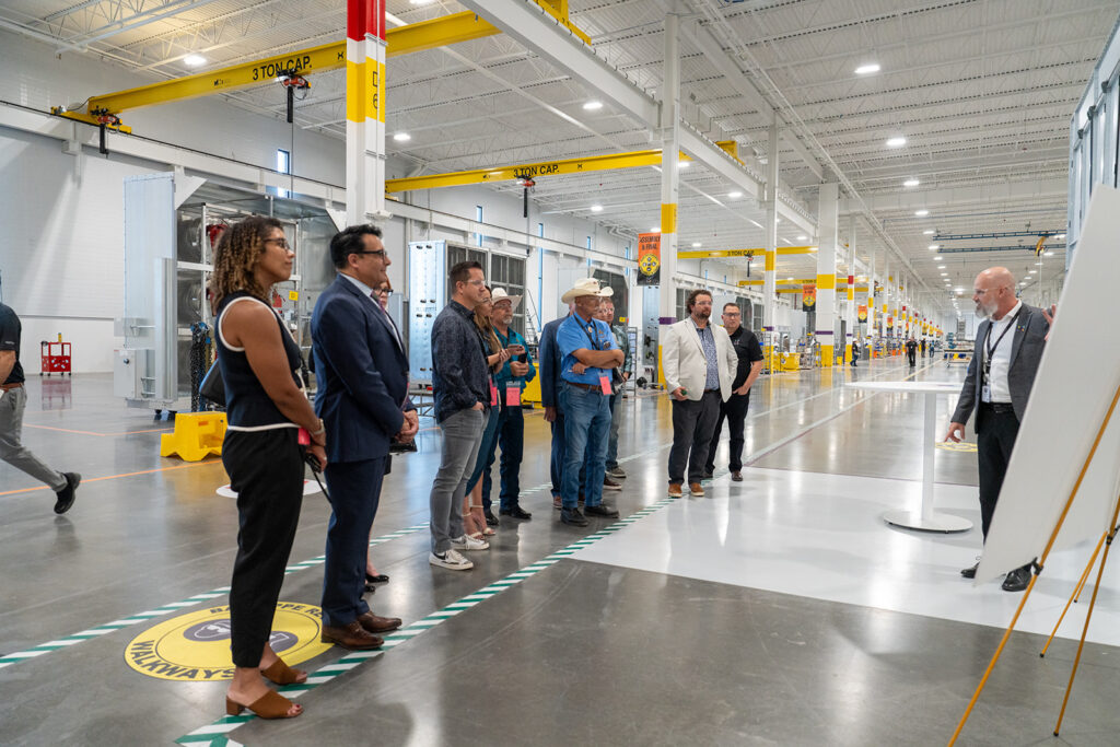 Ribbon cutting tour group listening to an XNRGY presenter on the production floor beneath overhead cranes at the Mesa Gateway facility.