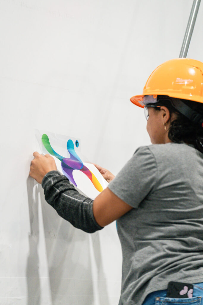 Worker in orange hard hat applying the colorful XNRGY X logo decal to a white interior wall at the Mesa Gateway facility.