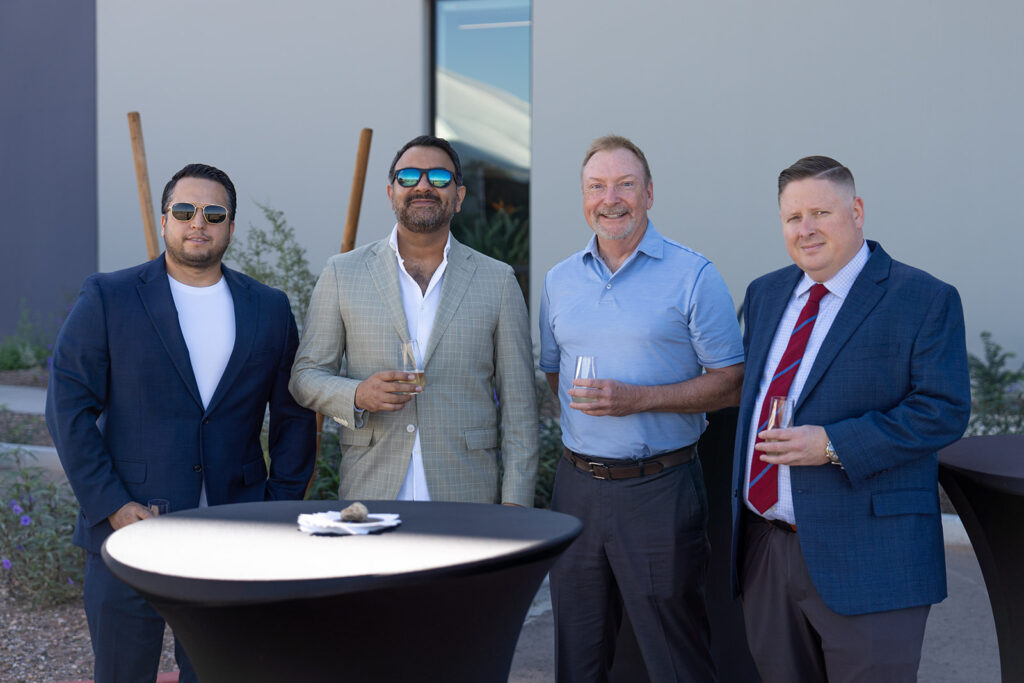 Four guests in business attire pose with champagne glasses at a cocktail table outside the XNRGY Mesa Gateway facility during the ribbon cutting welcome reception.