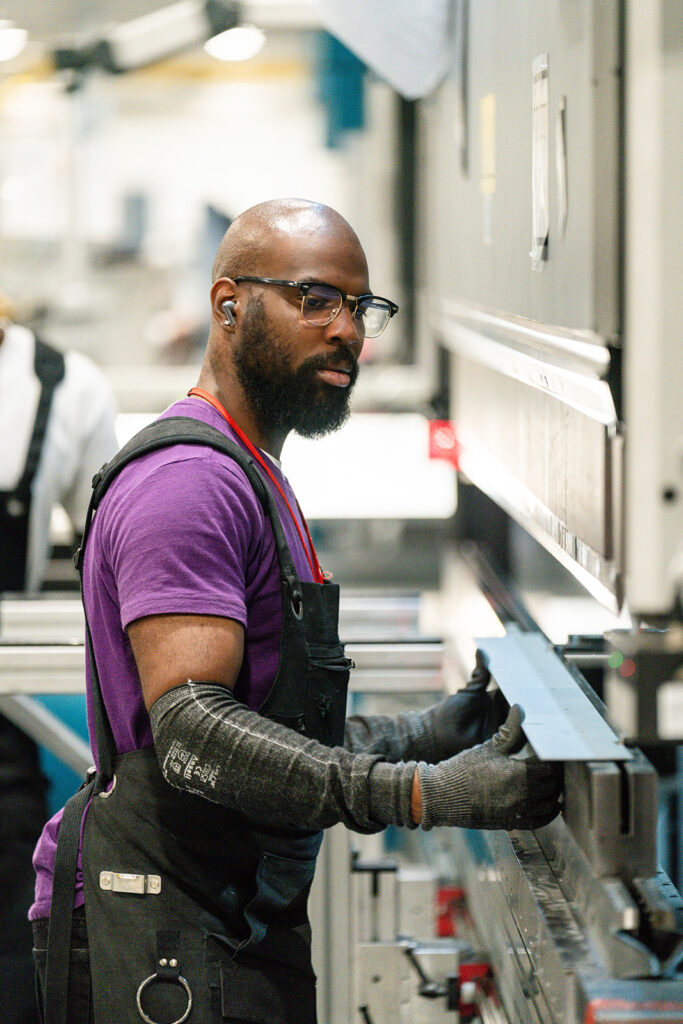 XNRGY worker in safety glasses operating a sheet metal fabrication machine at the Mesa Gateway facility.