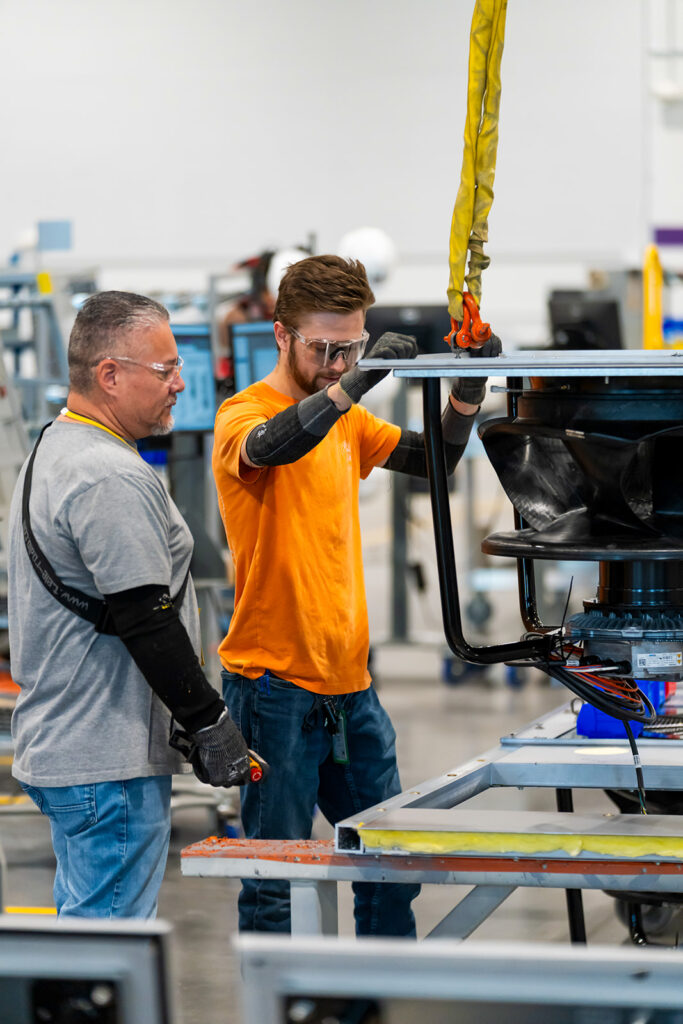 Two XNRGY production workers in safety glasses assembling a large cooling component on the factory floor at the Mesa Gateway facility.