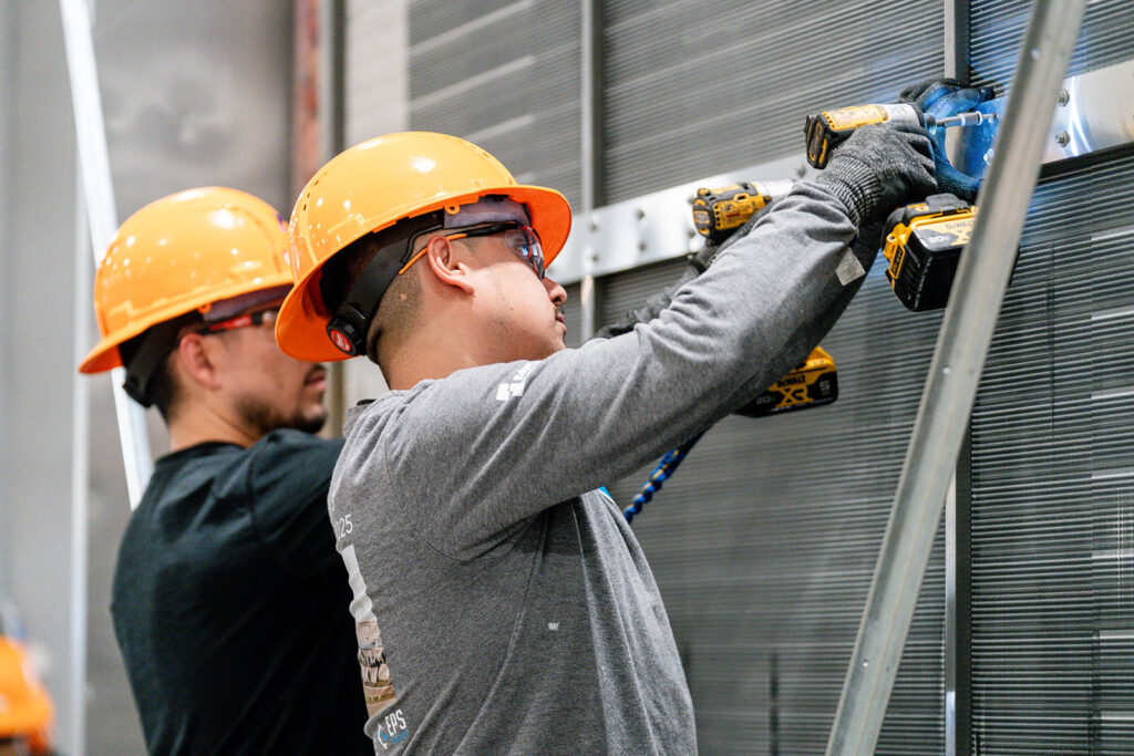 Two XNRGY team members in hard hats and safety glasses using cordless drills to install a component on a metal panel at the Mesa Gateway facility.