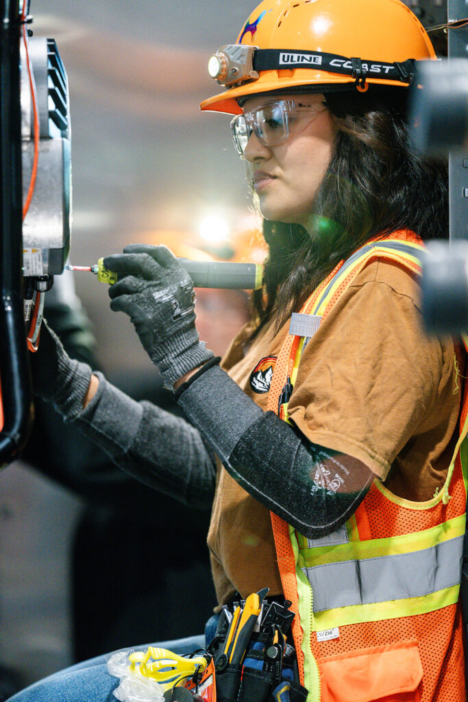 XNRGY technician in a hard hat with headlamp using a screwdriver inside an equipment cabinet at the Mesa Gateway facility.